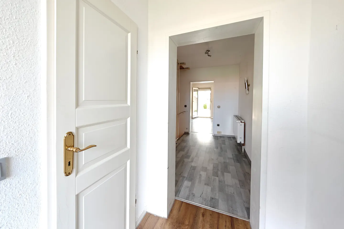 A bright, white doorway with a gold handle leads to a room with gray wood floors and a white radiator.