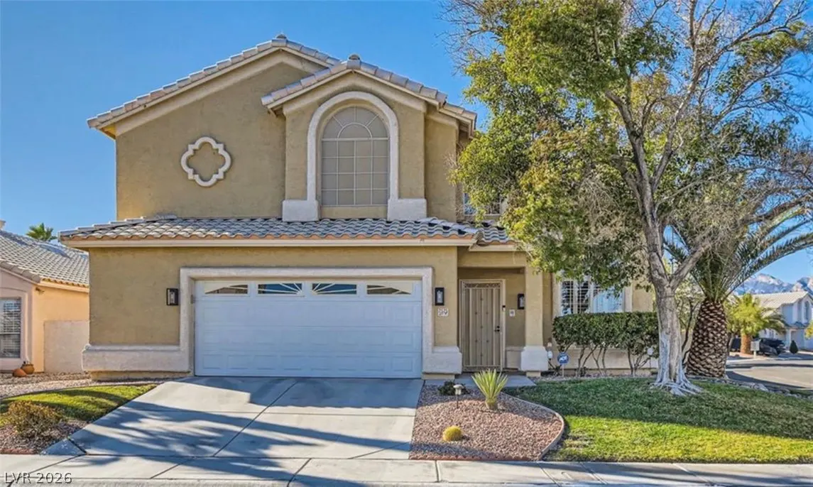 Two-story tan stucco suburban home with a white garage door, arched window, and a front yard with a tree and desert plants.