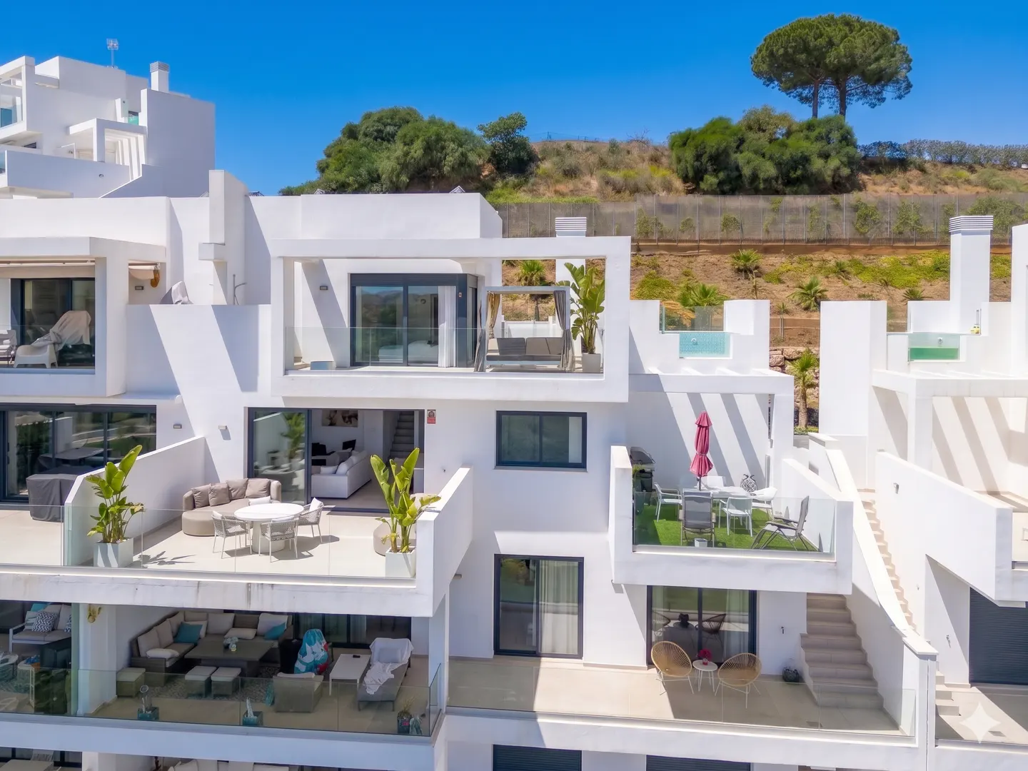 Modern white multi-story building with balconies and outdoor furniture under a clear blue sky. Hillside with trees in the background.