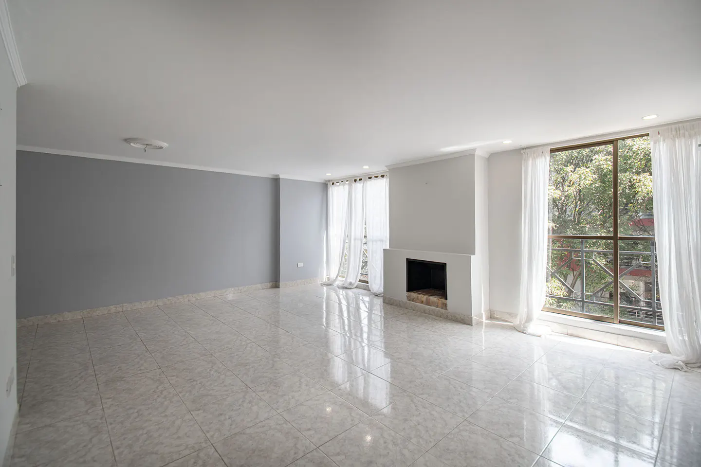 Empty living room with gray wall, tile floor, fireplace, and large windows with white curtains.