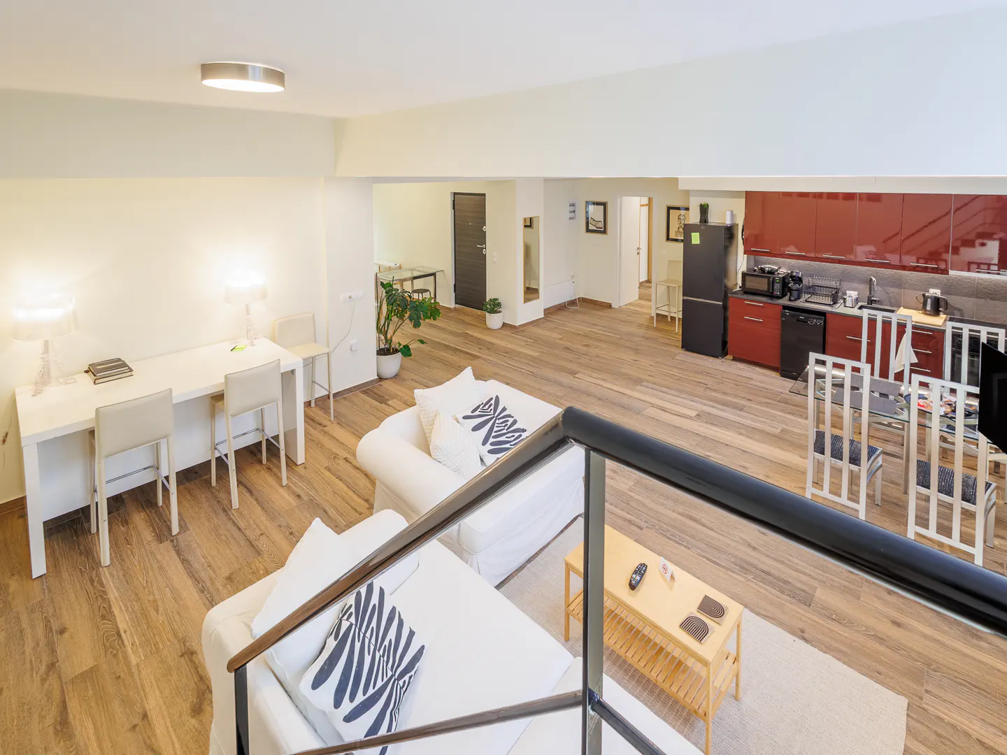Open-concept living space with wood floors, white sofas with patterned pillows, and a desk area. Red kitchen cabinets in the background.