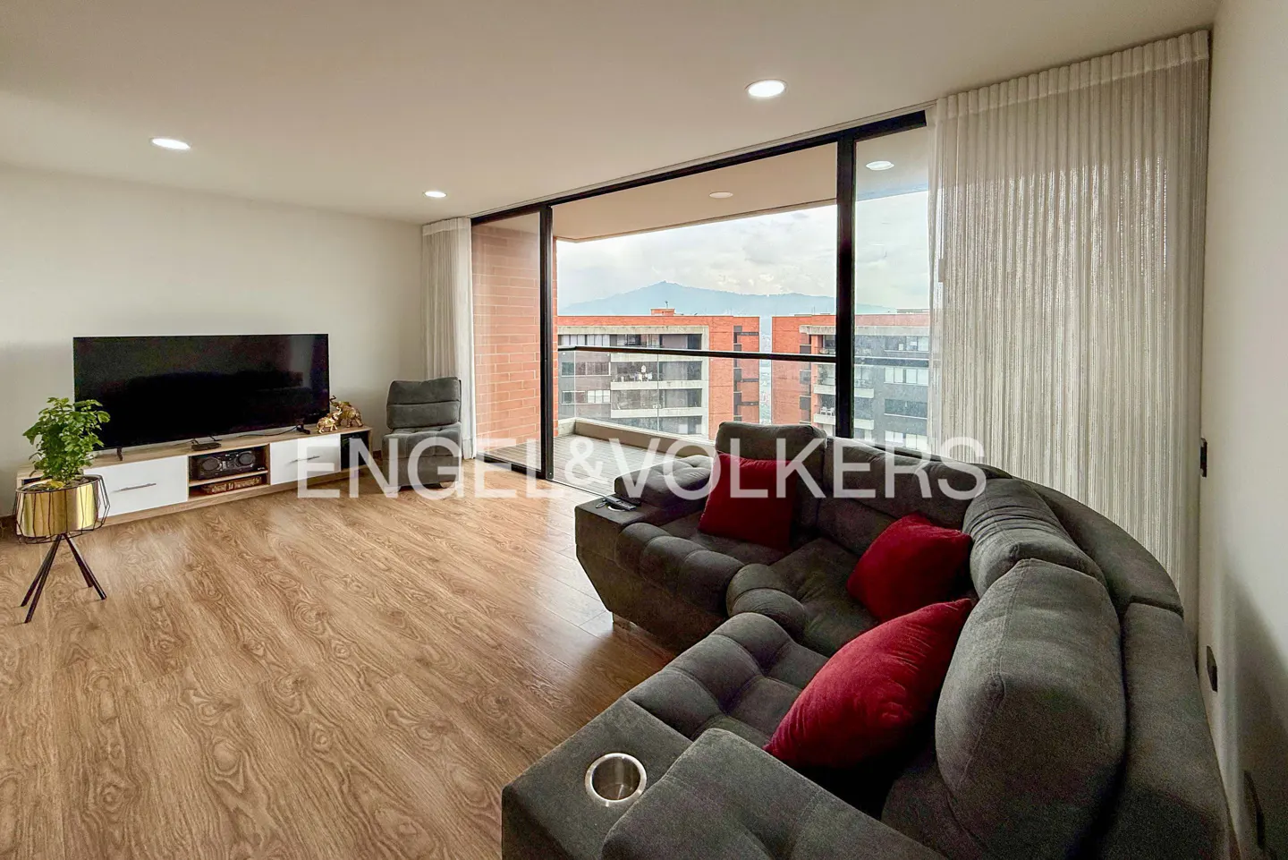 Modern living room with wood floors, grey sectional sofa with red pillows, TV console, and balcony with city mountain view.
