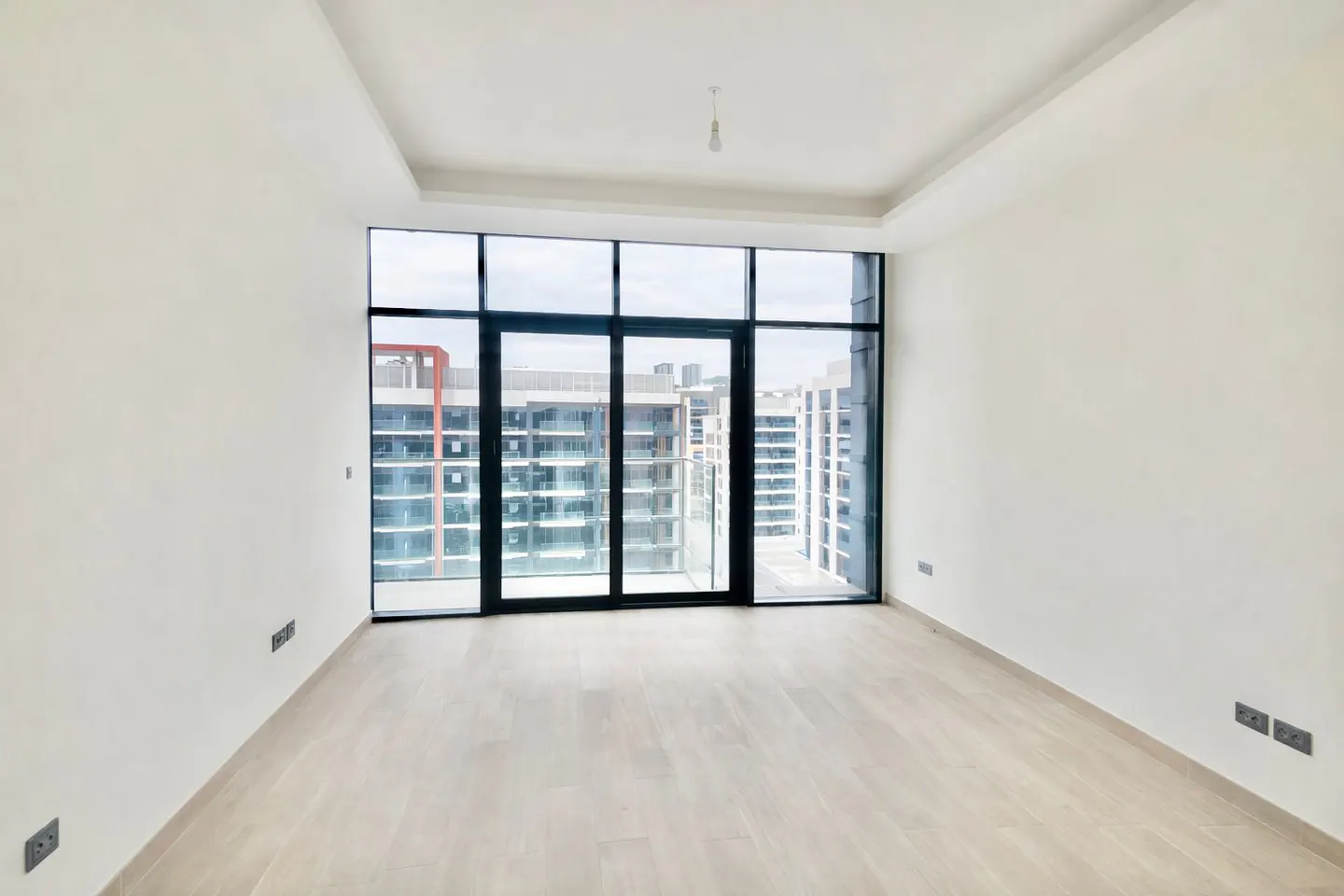 Bright, empty room with light wood floors and white walls. Large black-framed windows offer a city view.