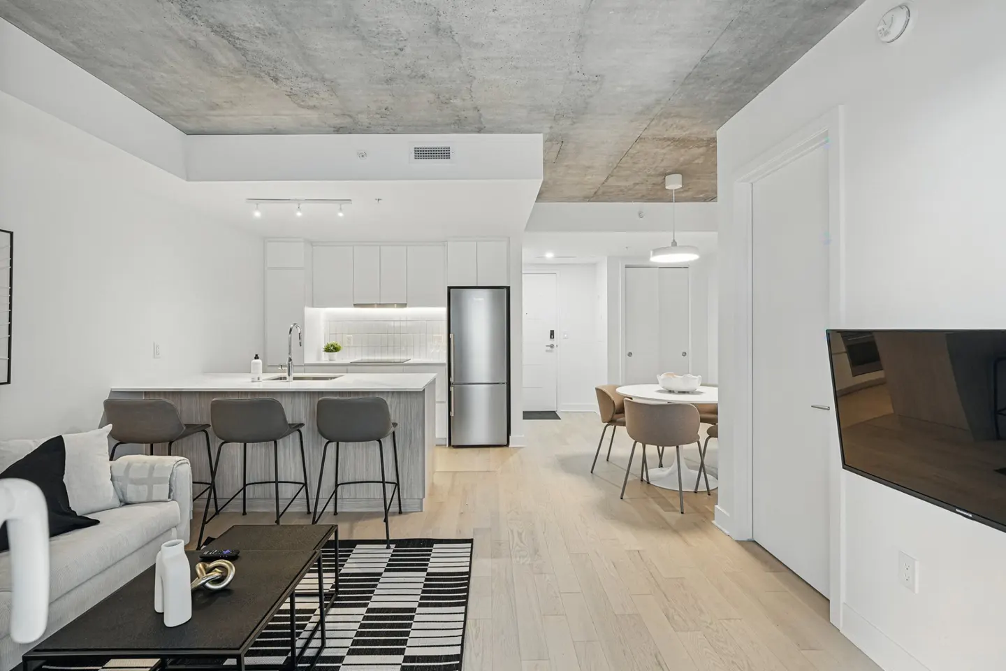 Bright, modern apartment interior with white walls, concrete ceiling, and light wood floors. Kitchen island with bar stools, dining table, and living area with sofa.