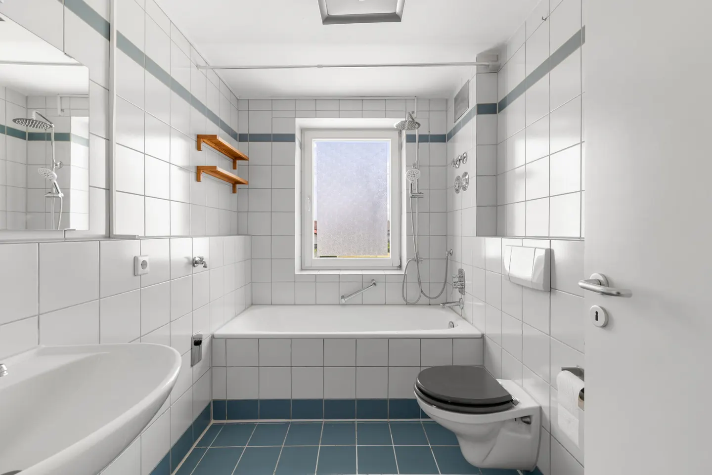Bright bathroom with white tiled walls, blue floor tiles, a bathtub under a window, a toilet, and a sink.