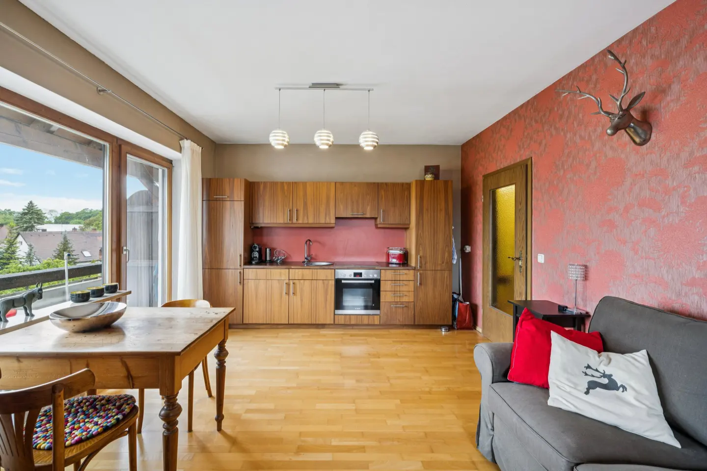 Open-plan kitchen and living area with wooden cabinets, a dining table, and a grey sofa against a textured red wall.