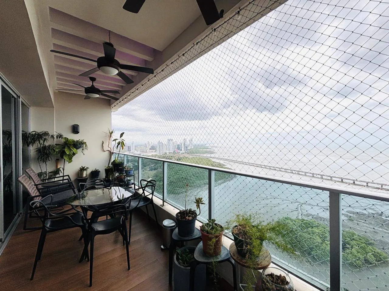 Balcony with black table and chairs, potted plants, and a safety net overlooking a city skyline and ocean view.