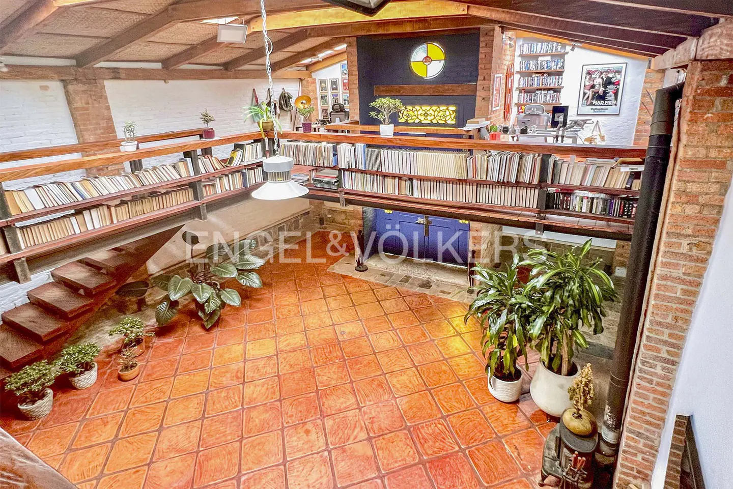 High-angle view of a rustic living room with terracotta tile floors, a wooden mezzanine library, and several potted plants.