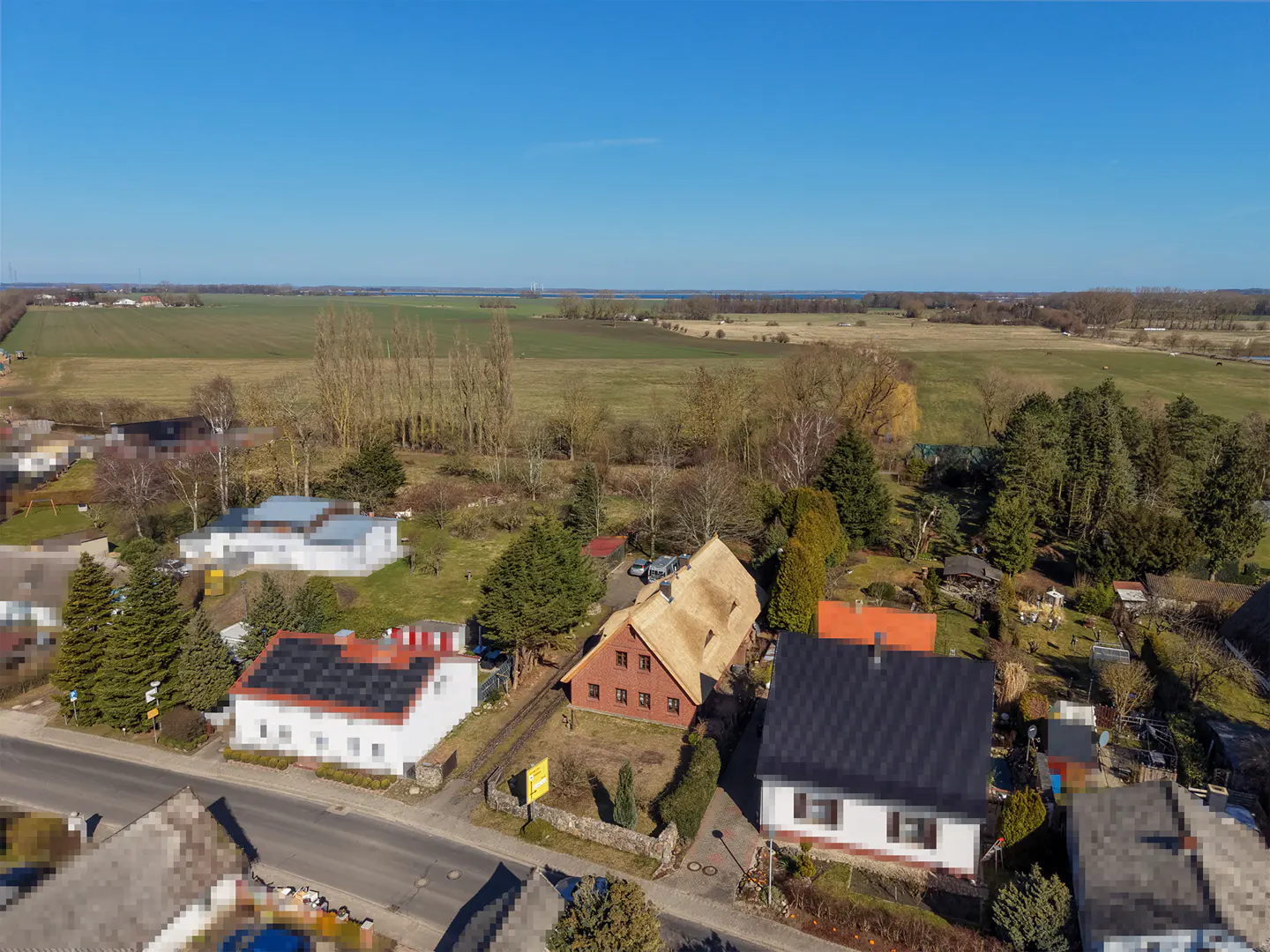 Aerial view of a rural neighborhood with houses, trees, and a large field under a clear blue sky. One house has a distinctive thatched roof.