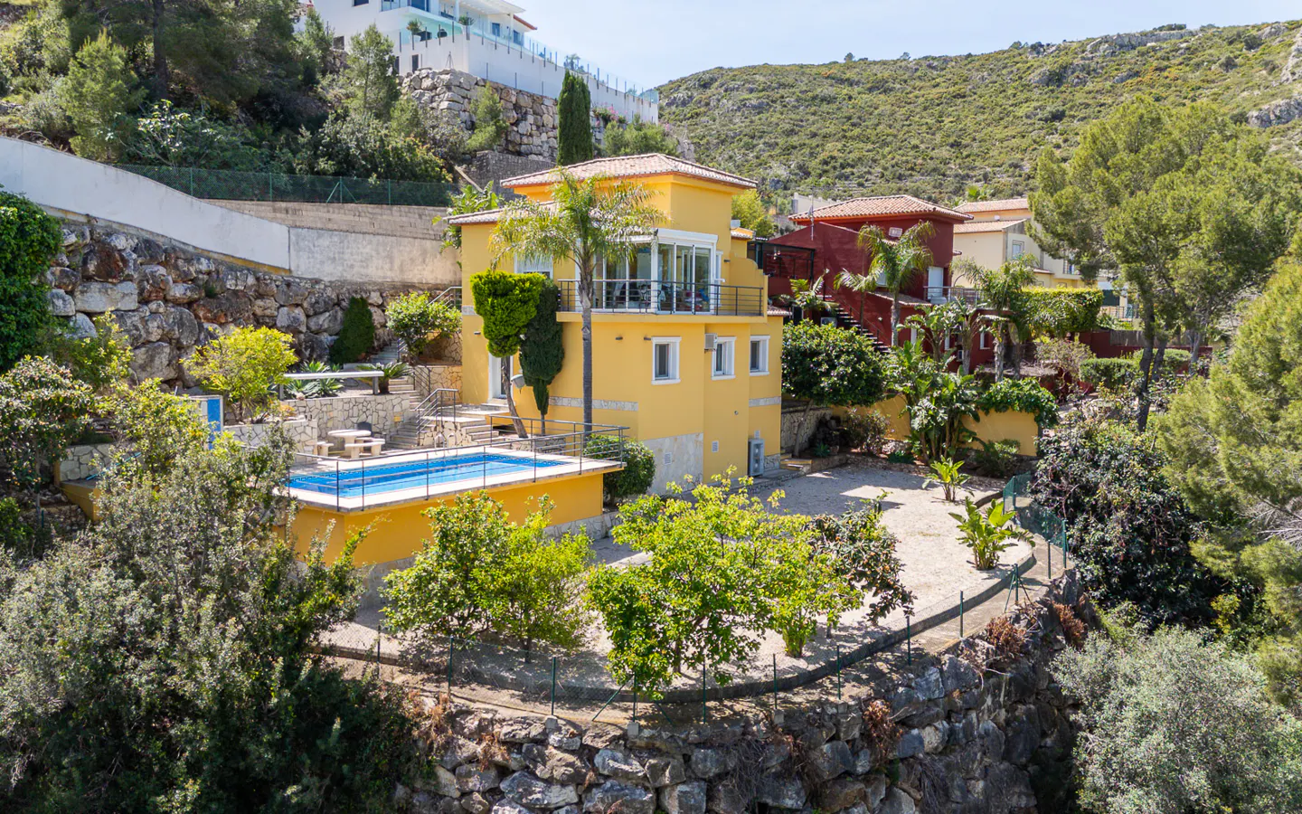 A yellow house with a pool sits on a rocky hillside surrounded by green trees and other houses.