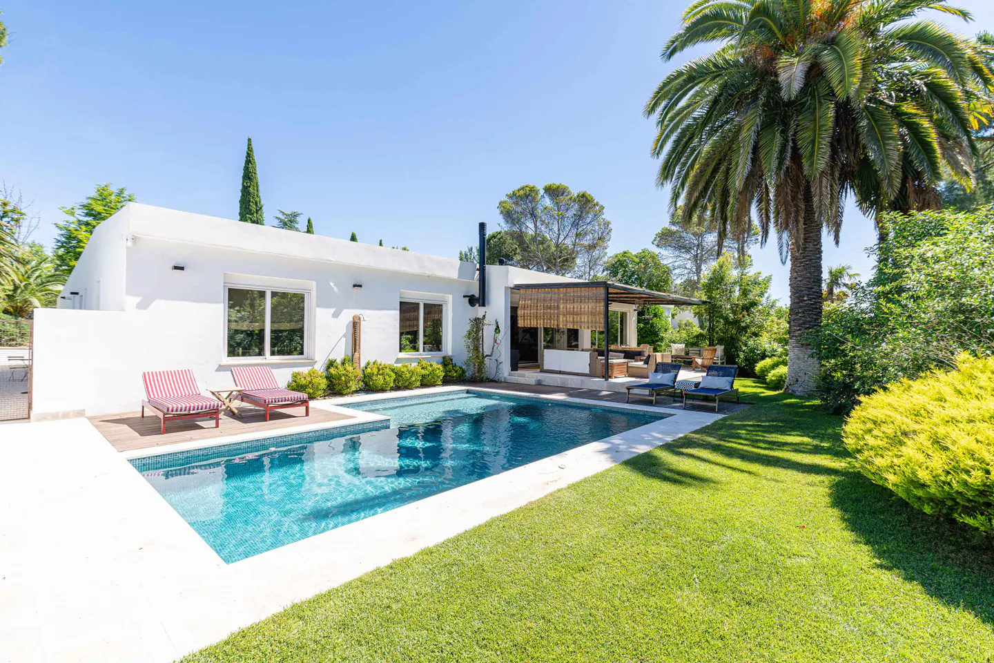 White modern villa with a swimming pool, lounge chairs, and a lush garden under a clear blue sky.