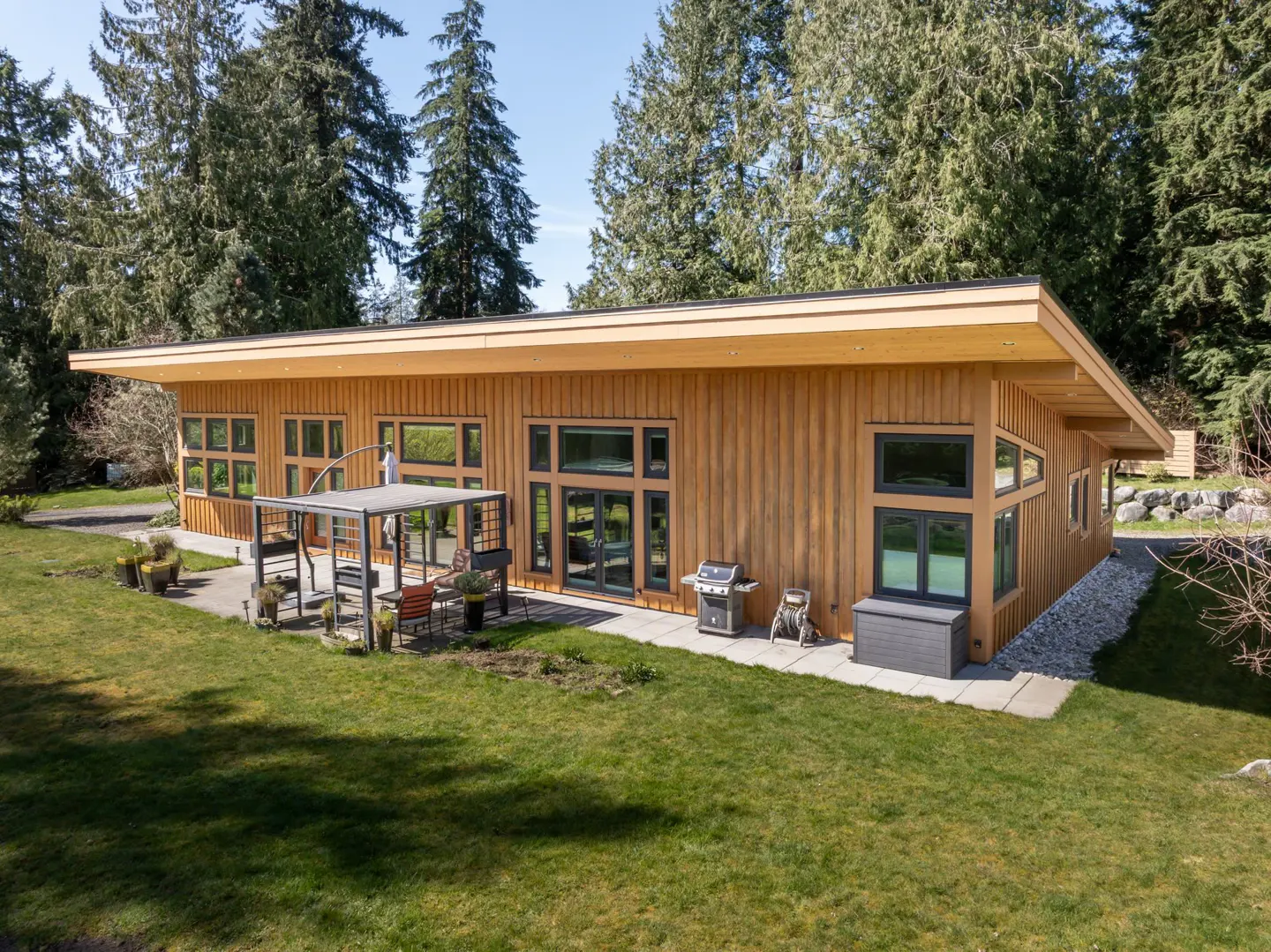 Exterior view of a modern, single-story home with wood siding, black framed windows, and a patio with outdoor furniture. Tall trees surround the property.