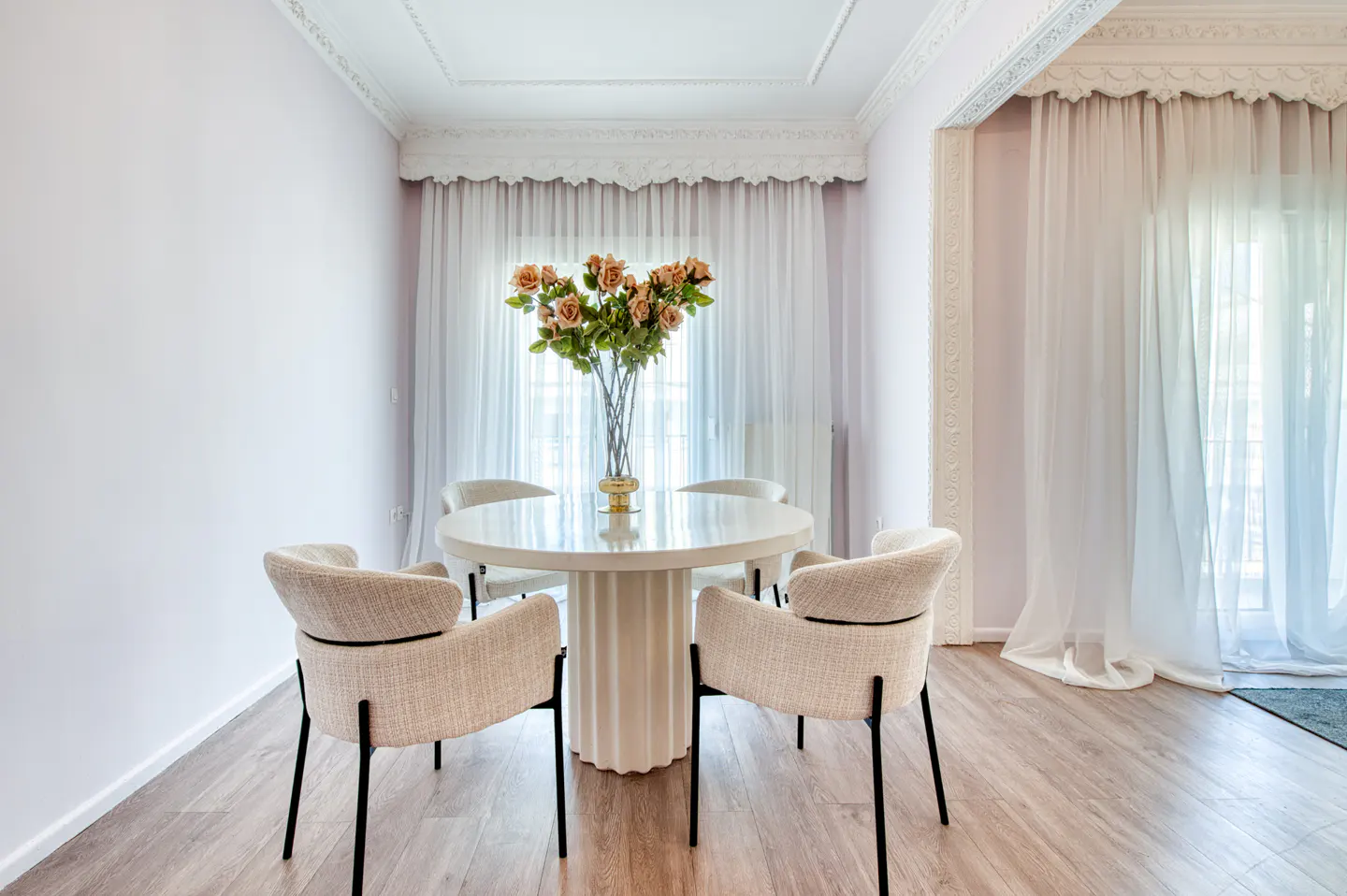 Dining room with a round white table, four beige chairs, and a vase of roses. White walls, wood floors, and sheer curtains.