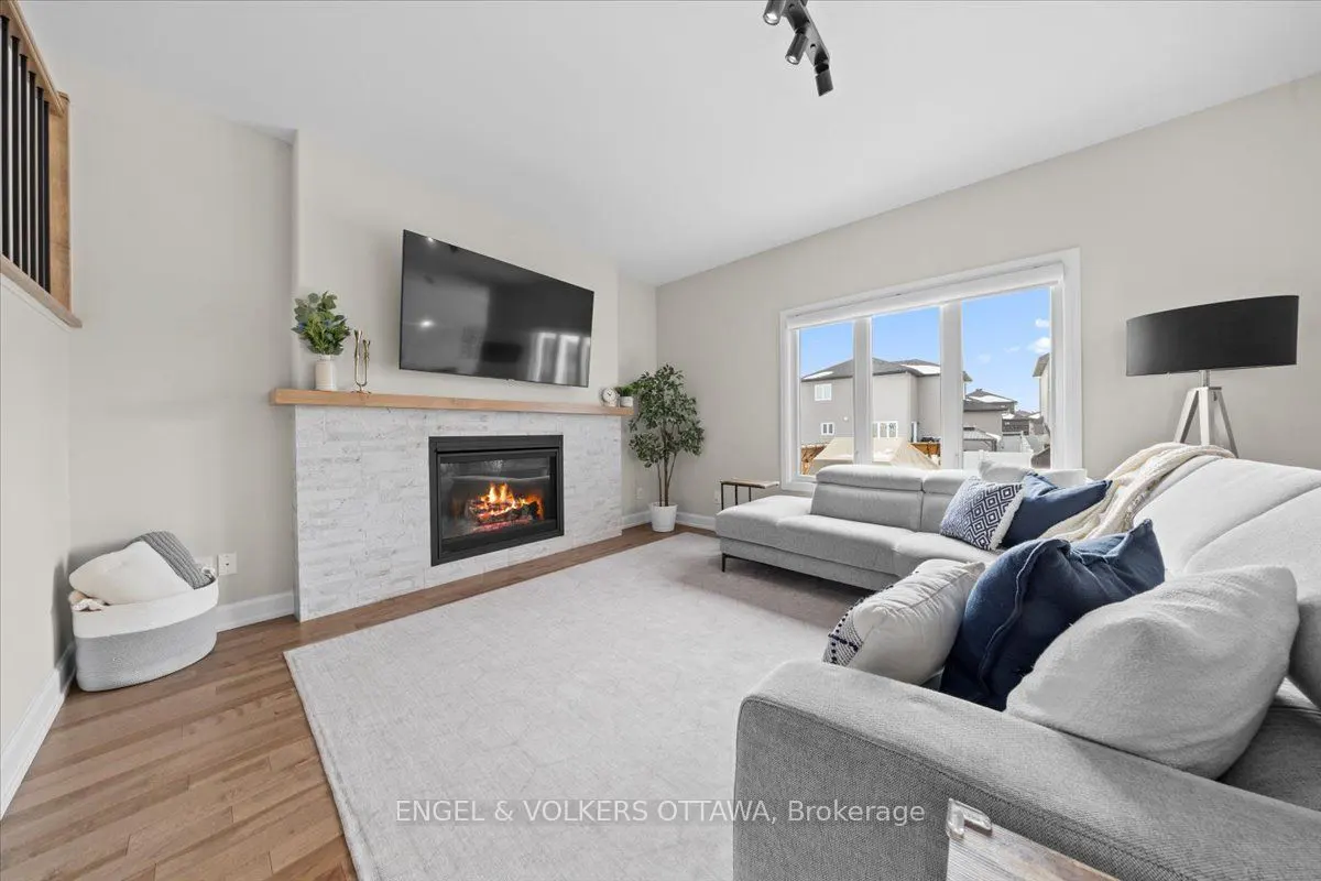 Bright living room with a gray sectional sofa, blue pillows, and a white brick fireplace with a TV above. A large window overlooks a suburban street.