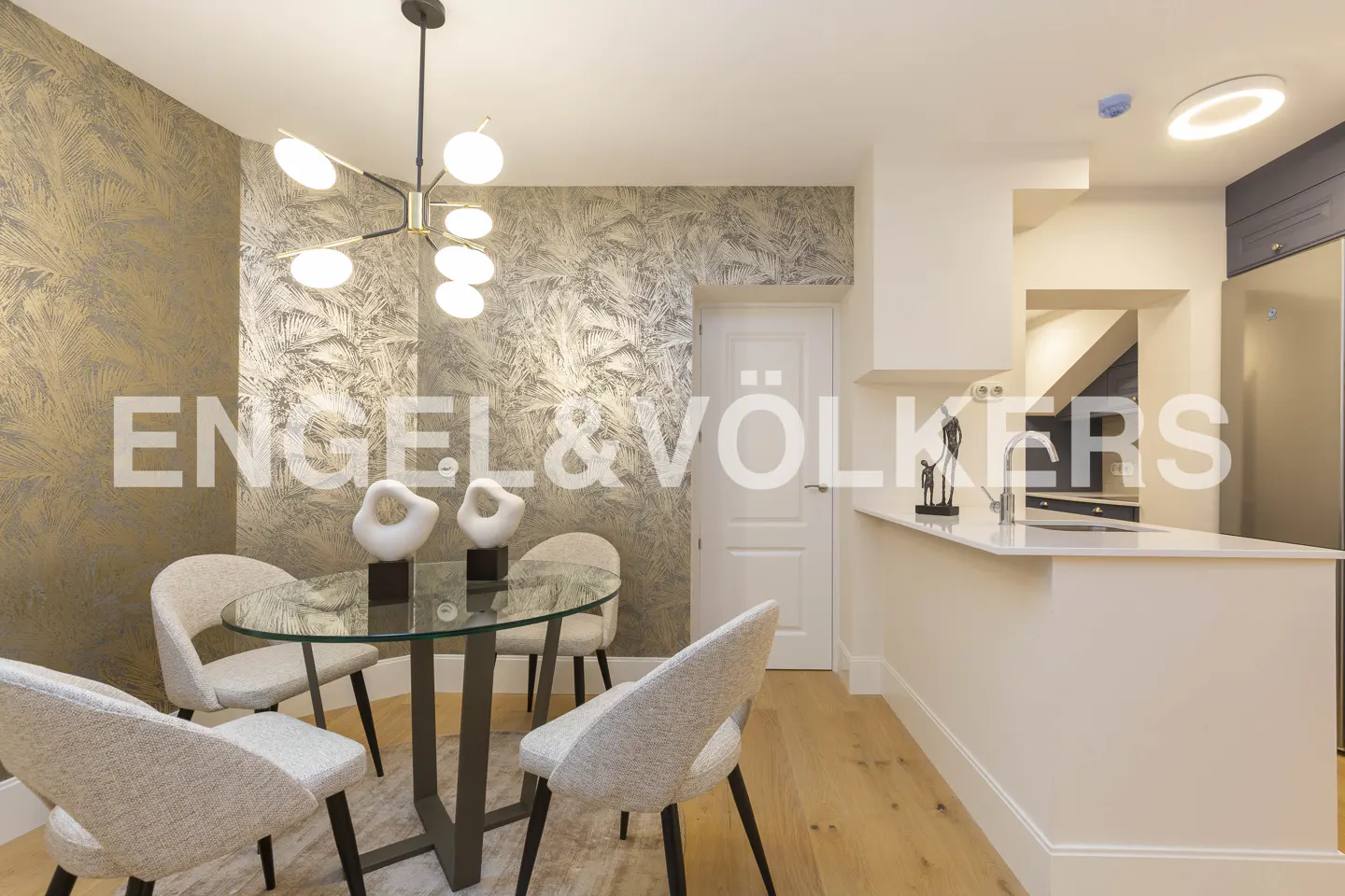 Modern dining area with a glass table, four light-colored chairs, a unique chandelier, and metallic palm-leaf wallpaper.