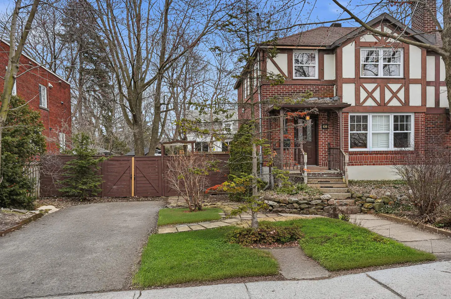 A two-story brick house with white trim and a brown roof, set back from the street with a stone walkway and green lawn. A brown fence is on the left.