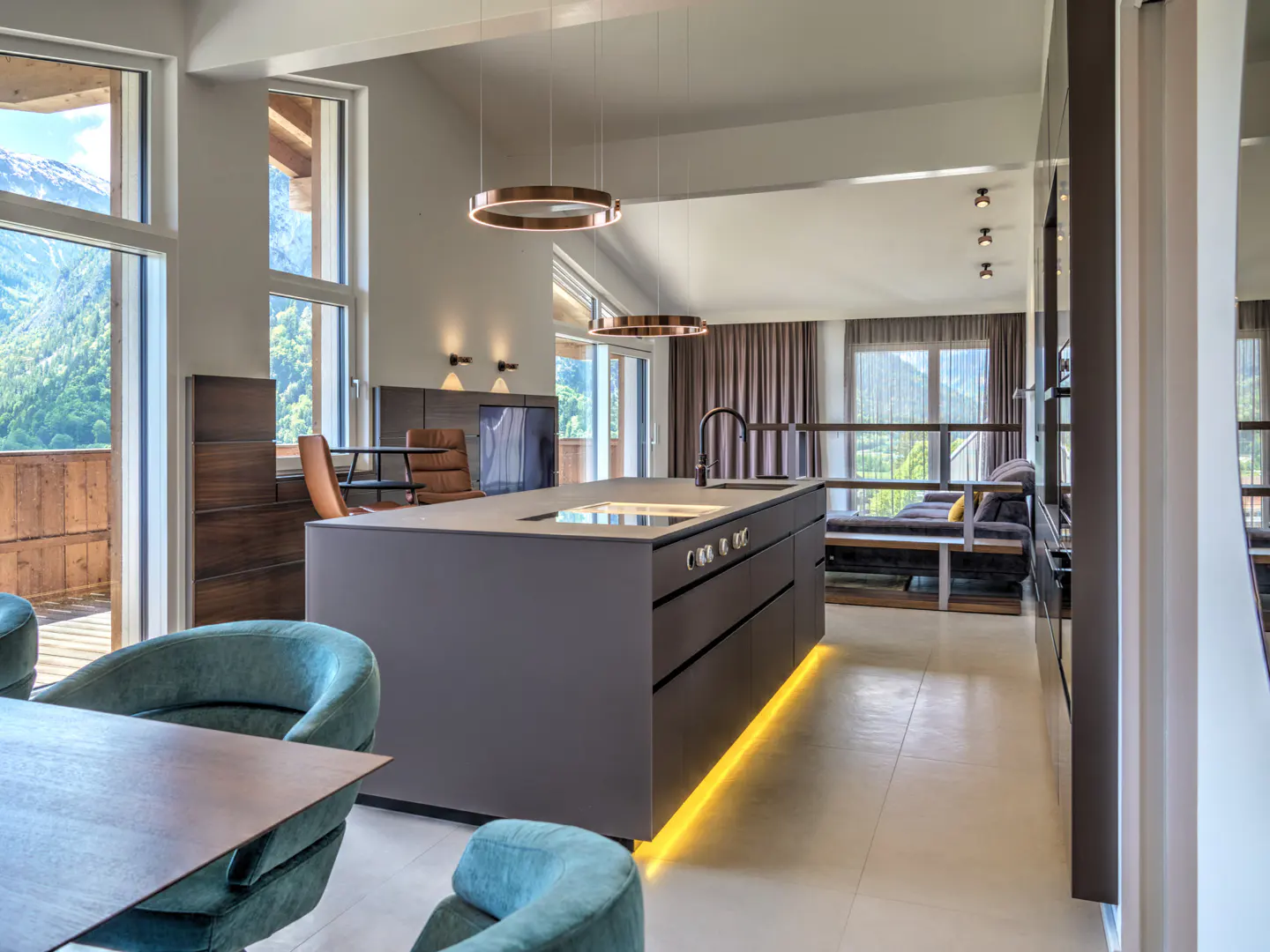Modern kitchen with a dark gray island, under-cabinet lighting, teal chairs, and large windows overlooking mountains.