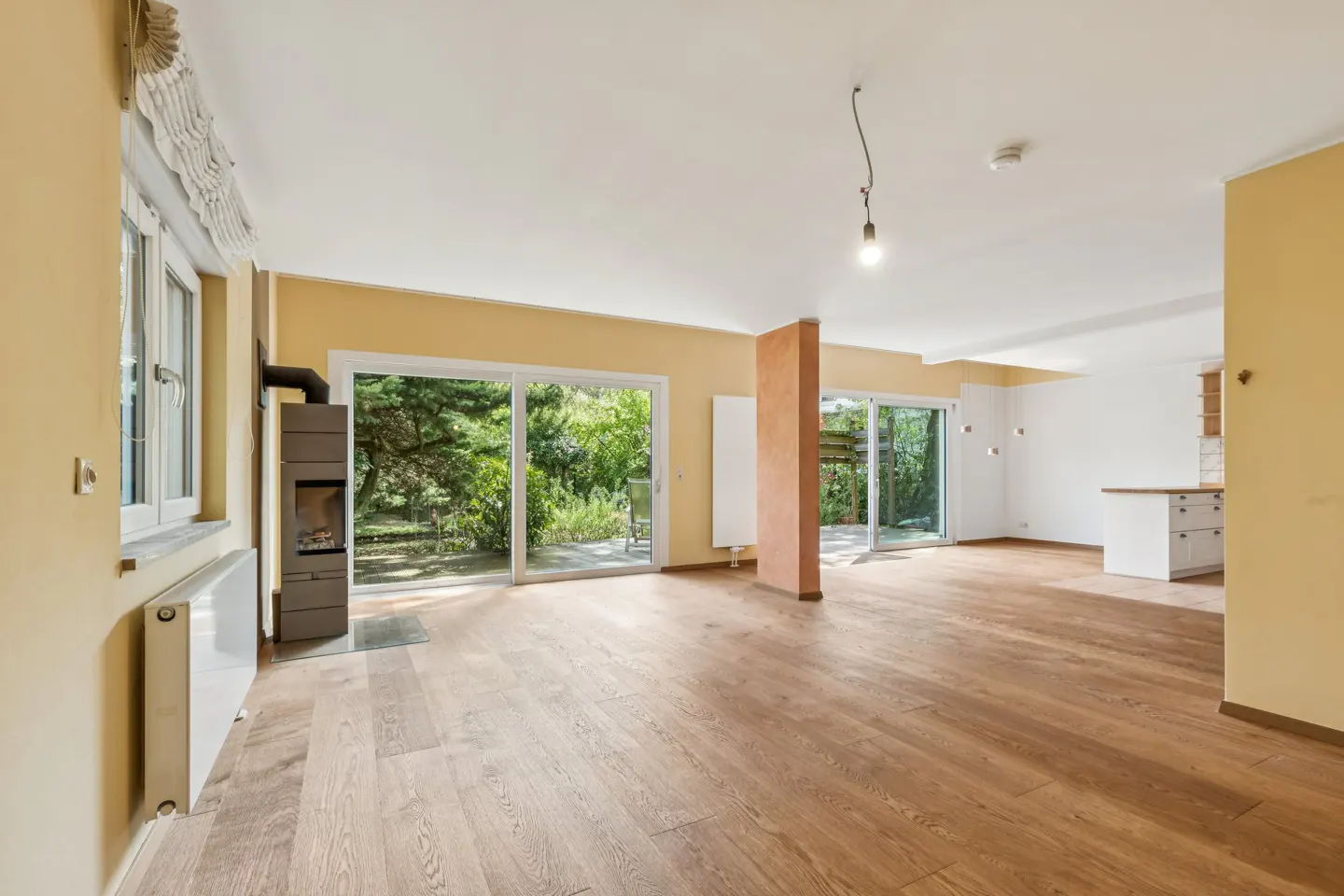 Bright, empty living room with wood floors, yellow walls, and sliding glass doors to a green yard. A modern fireplace stands near a window.