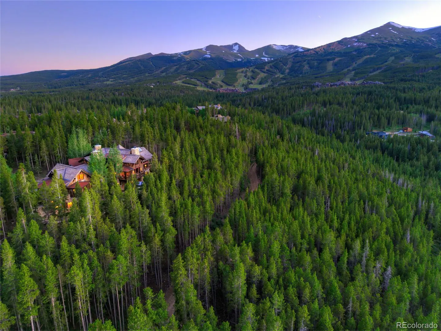 Aerial view of a large, wood-clad house nestled in a dense green forest with snow-capped mountains in the background.