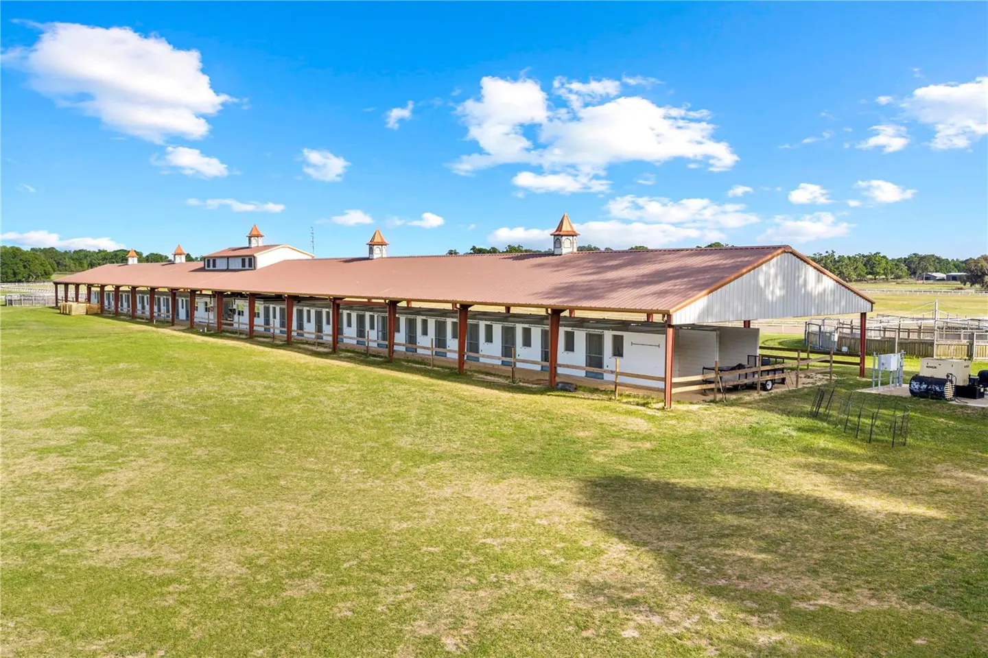 Exterior view of a horse stable with a red roof and white walls, surrounded by a green field under a blue sky with clouds.