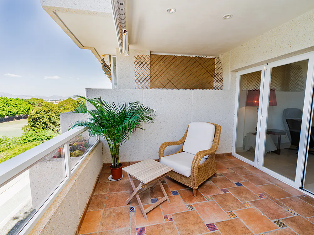 A sunny balcony with a wicker chair, small table, and potted palm. The floor is tiled in terracotta, and a view of trees is visible.