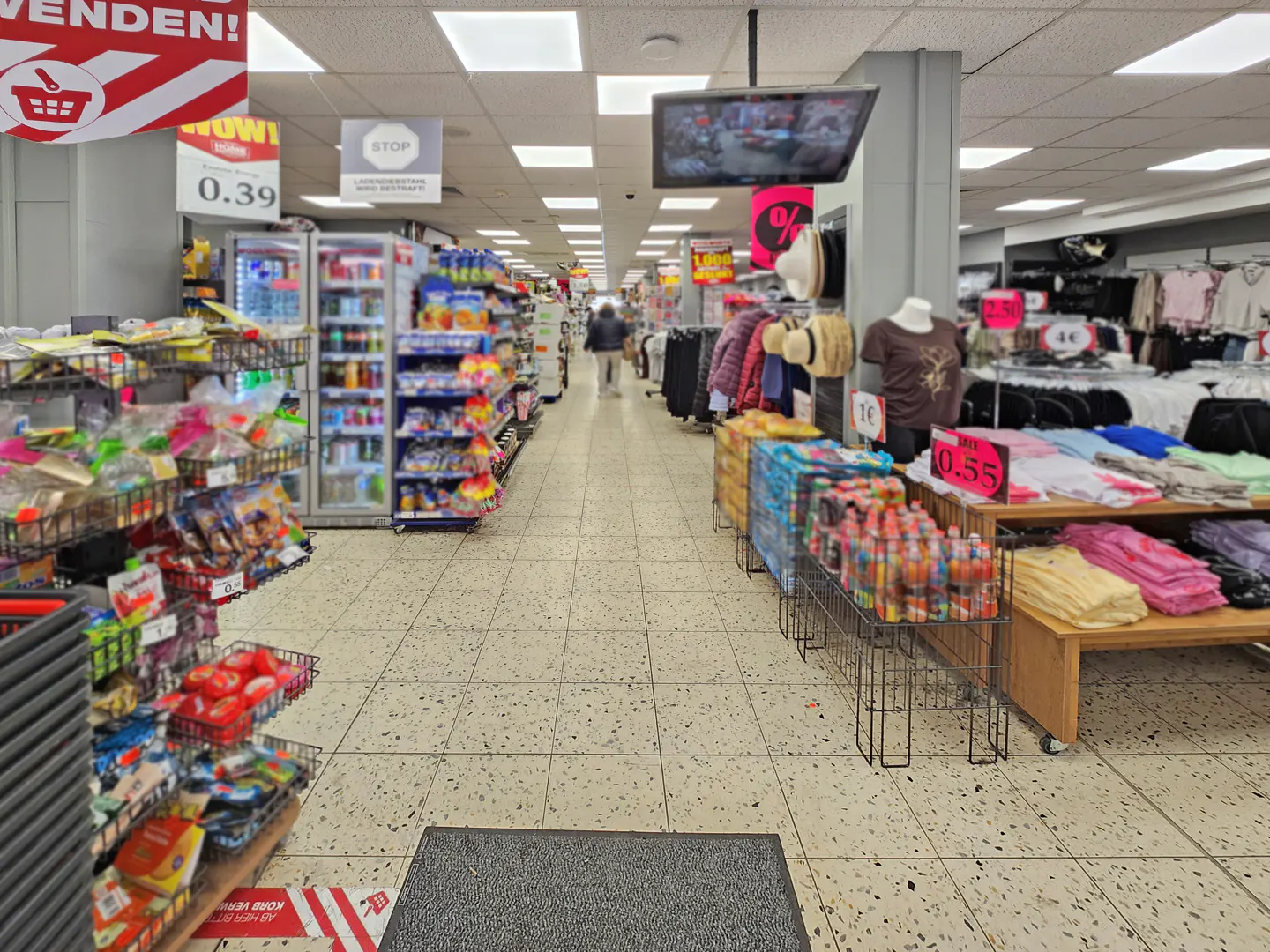 Aisles of a grocery store with shelves of food and clothing on display. A TV hangs from the ceiling.