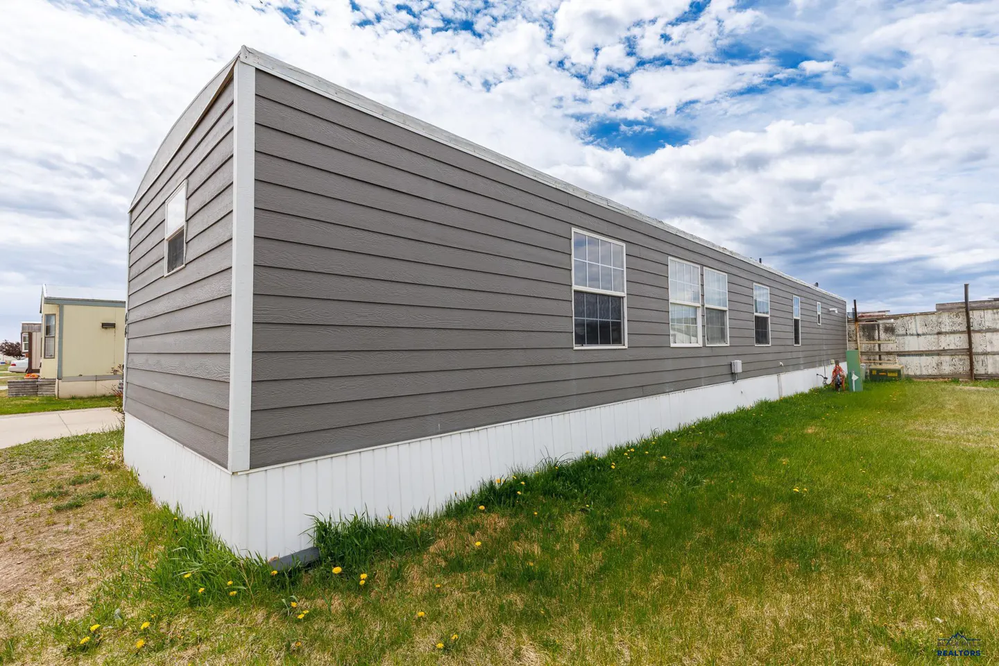 Side view of a long, grey manufactured home with white skirting, set on a grassy lot under a cloudy blue sky.