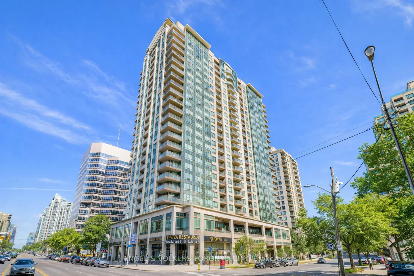 Exterior view of a tall, modern condo building with glass balconies, street-level retail, and cars parked along the street.