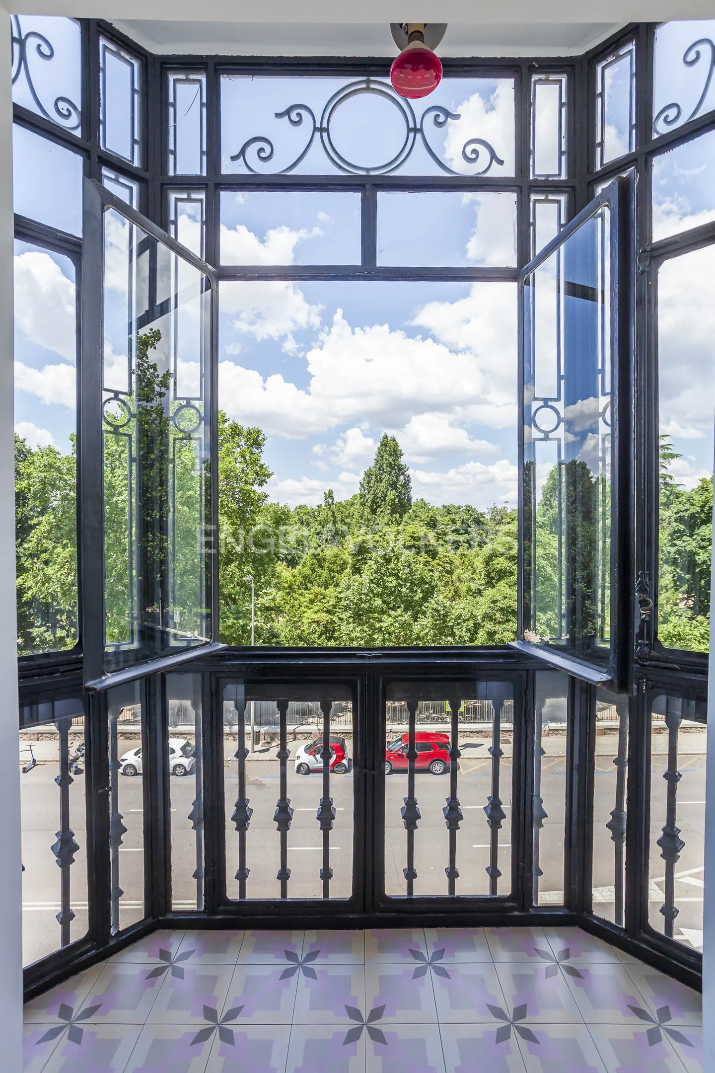 View from a black-framed bay window with open panes, overlooking a tree-lined street with cars and a tiled floor.