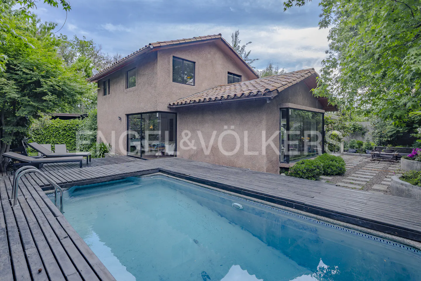 Two-story stucco house with a tiled roof, featuring a swimming pool and wooden deck in a lush, green backyard.