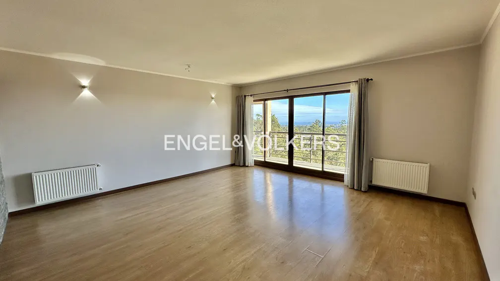 An empty room with wood floors, beige walls, and a large window with a view of trees and the ocean. Radiators are on the walls.