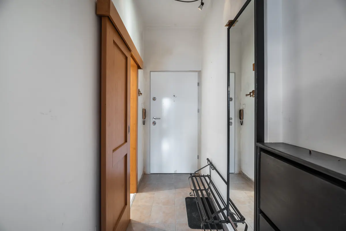 Hallway with a light wood sliding door, white walls, and a white front door. A black metal rack and shoe cabinet are visible.