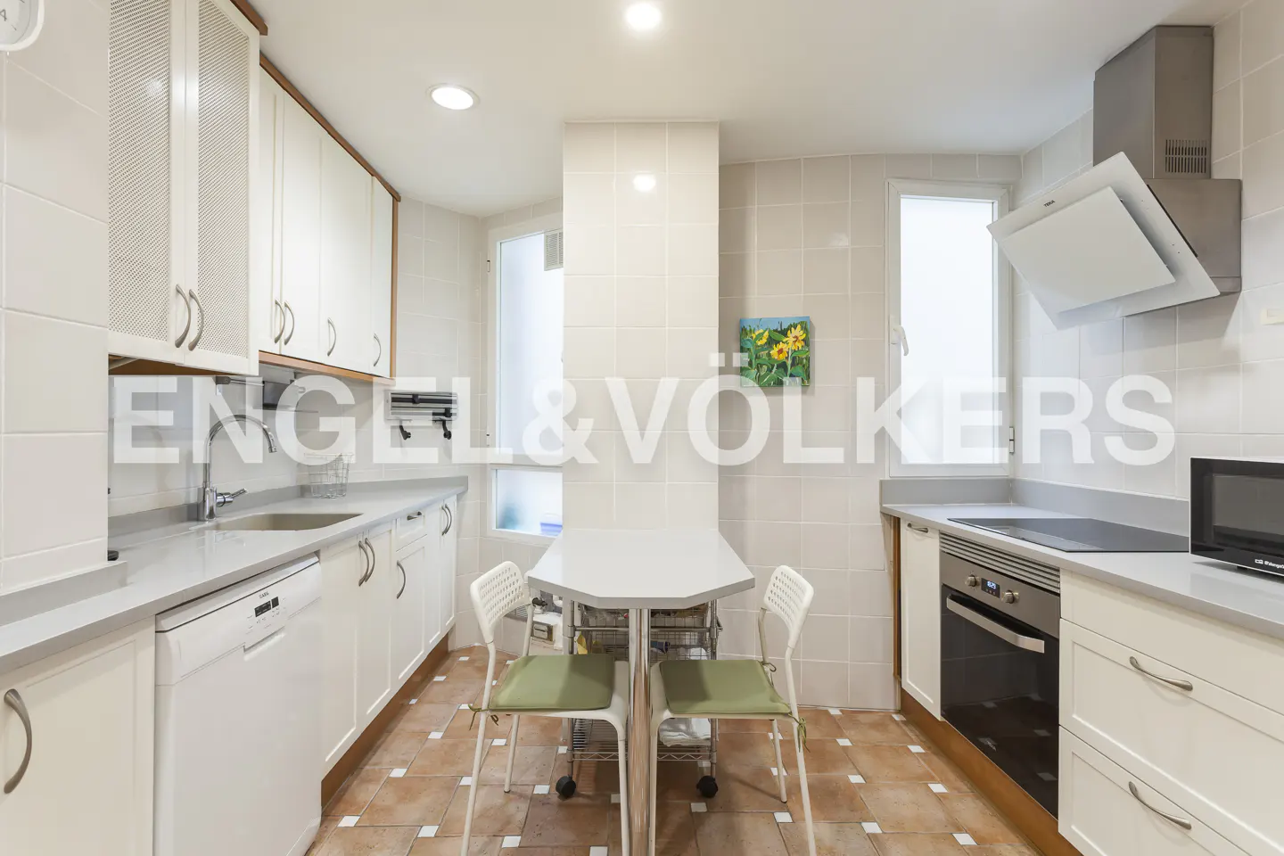 A bright kitchen with white cabinets, gray countertops, and a small white table with two chairs. The floor is tiled in brown and white.