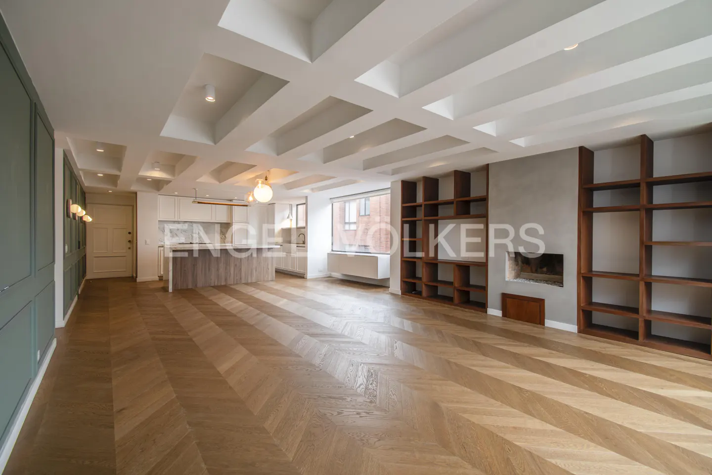 A wide shot of a modern apartment with herringbone wood floors, a white coffered ceiling, and a kitchen in the background.