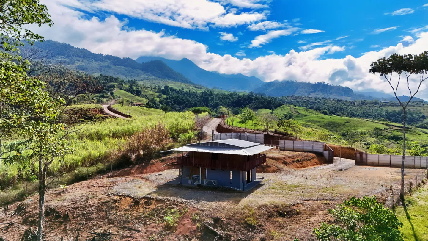 A two-story house sits on a hill with mountains in the background under a blue sky with white clouds.
