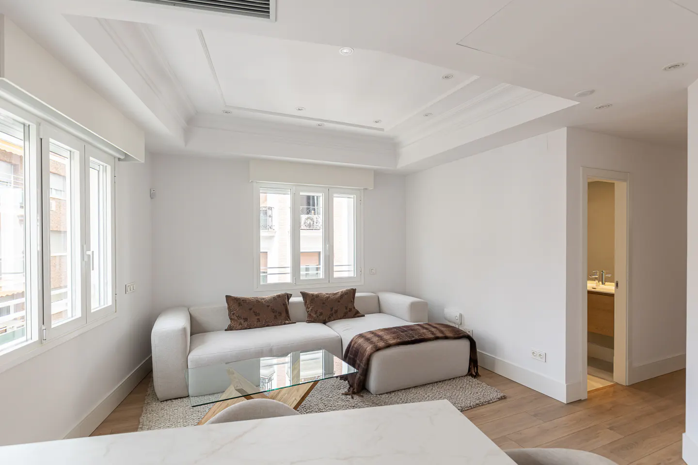 Bright living room with white walls, a white sofa with brown pillows, and a glass coffee table. Windows overlook a city view.