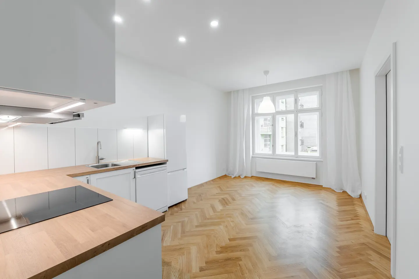 Bright, modern apartment with white walls, herringbone wood floors, and a white kitchen with wood countertops. A large window with white curtains lets in natural light.
