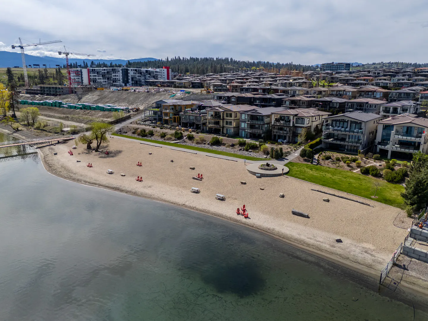 Aerial view of a sandy beach with red chairs, picnic tables, and lakefront homes under a cloudy sky.