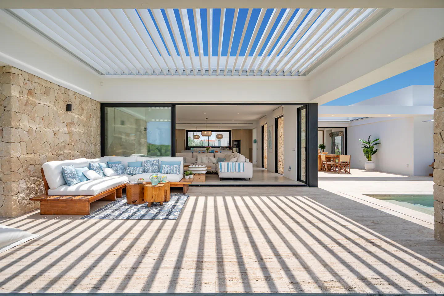 Modern patio with white sectional sofa, stone walls, and a louvered roof casting striped shadows on the tiled floor.