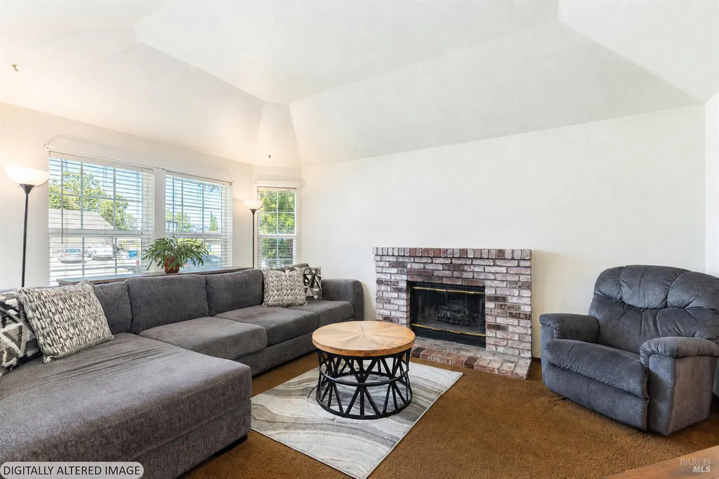 Bright living room with gray sectional, brick fireplace, and gray recliner. A round wood table sits on a gray and white rug.
