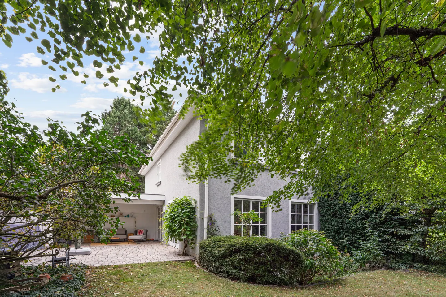 Exterior view of a two-story gray house with white trim, surrounded by green trees and lawn. A patio with furniture is visible.