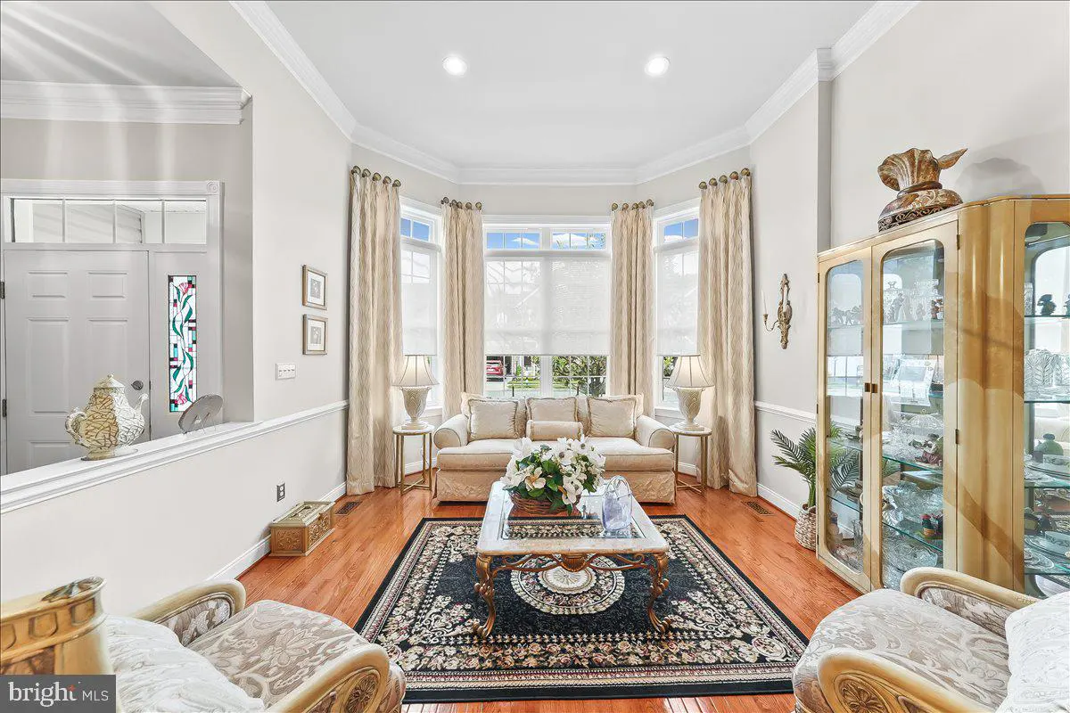 Bright living room with hardwood floors, beige sofa, and a large bay window with cream-colored curtains. A glass cabinet stands to the right.