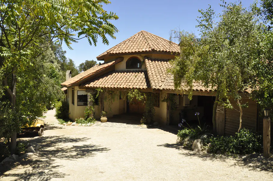 A tan house with a red tile roof is surrounded by trees and a gravel driveway.