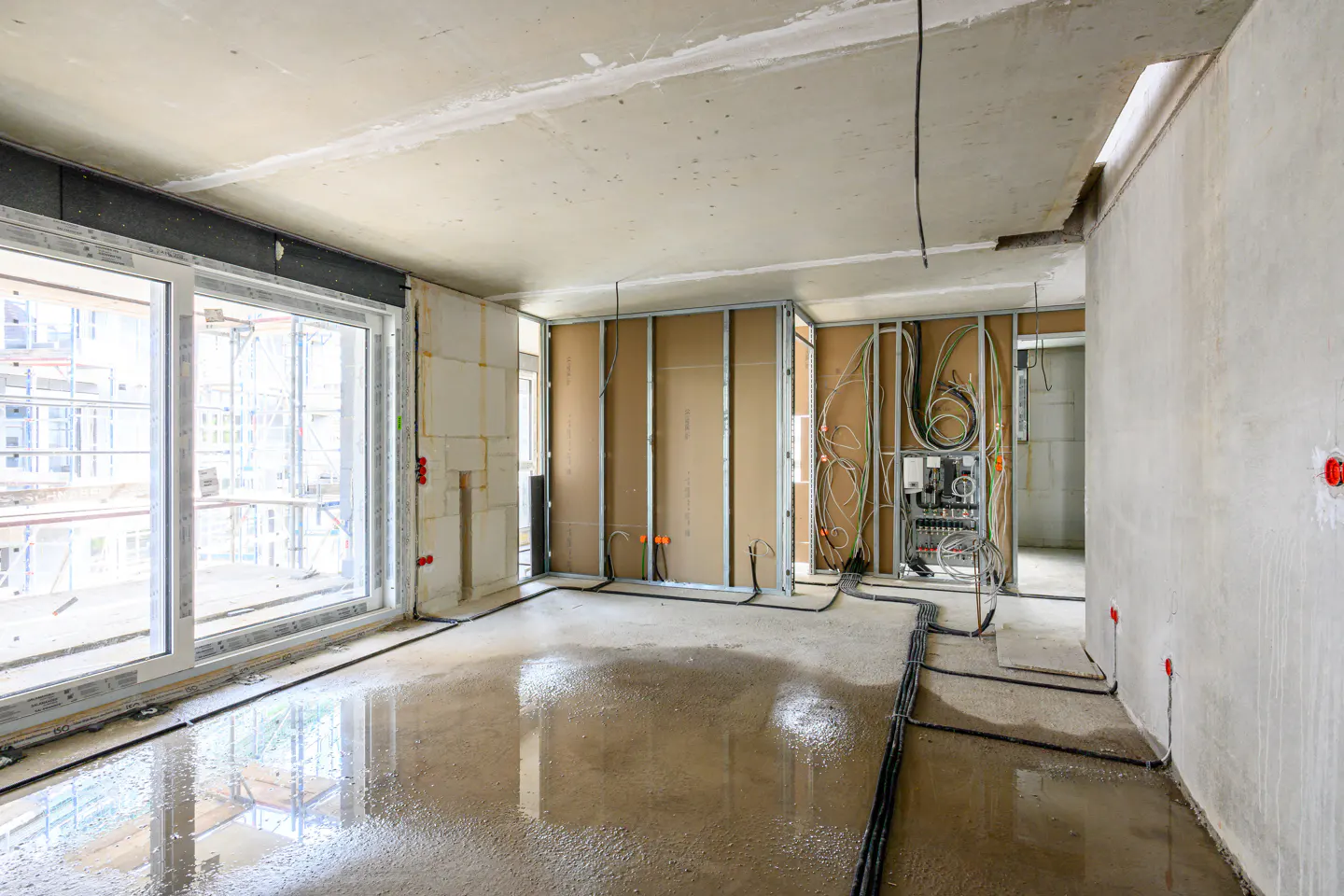 Interior view of a construction site with large windows, exposed wiring, and a wet concrete floor.