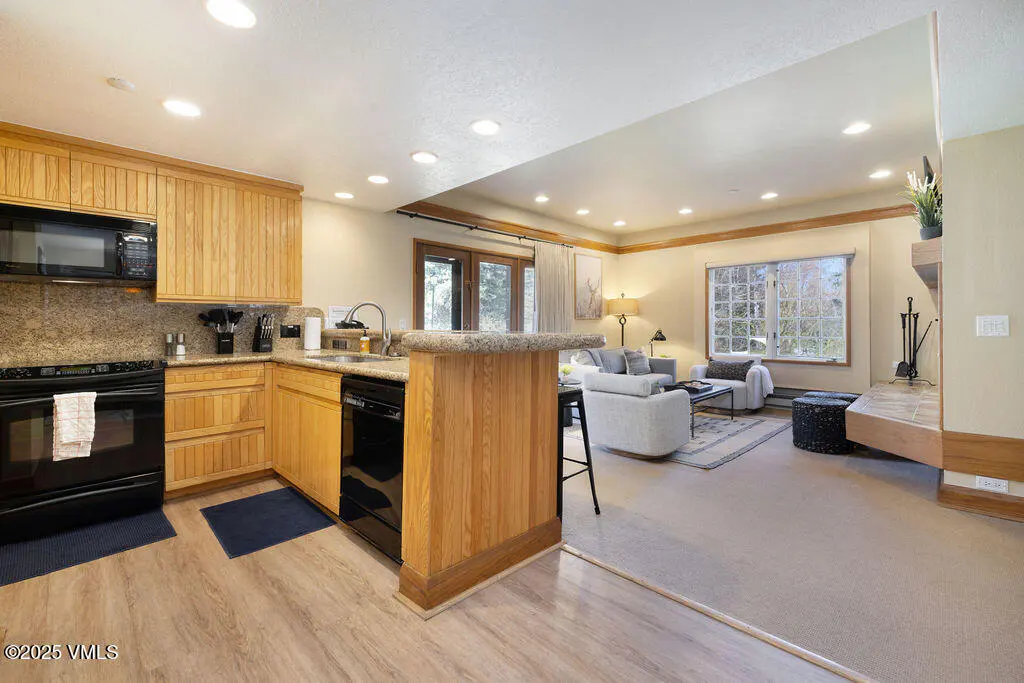 Open-concept living space with wood cabinets, black appliances, and a living room with a fireplace and gray sofa.