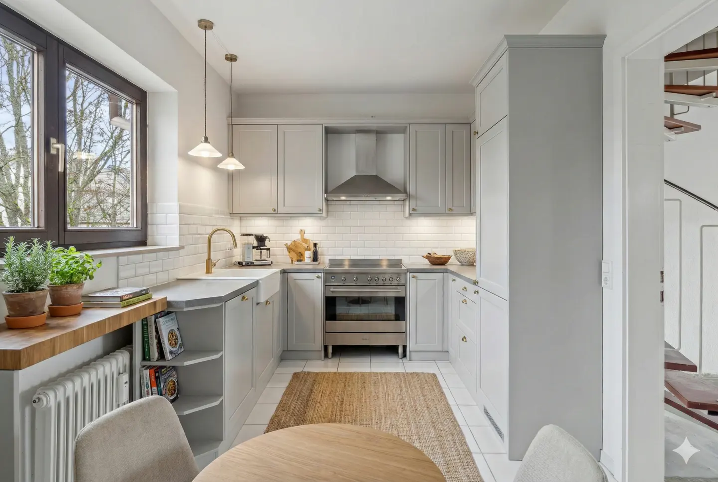 A bright kitchen with gray cabinets, white subway tile, and a jute rug. A round wooden table sits in the foreground. A window overlooks trees.