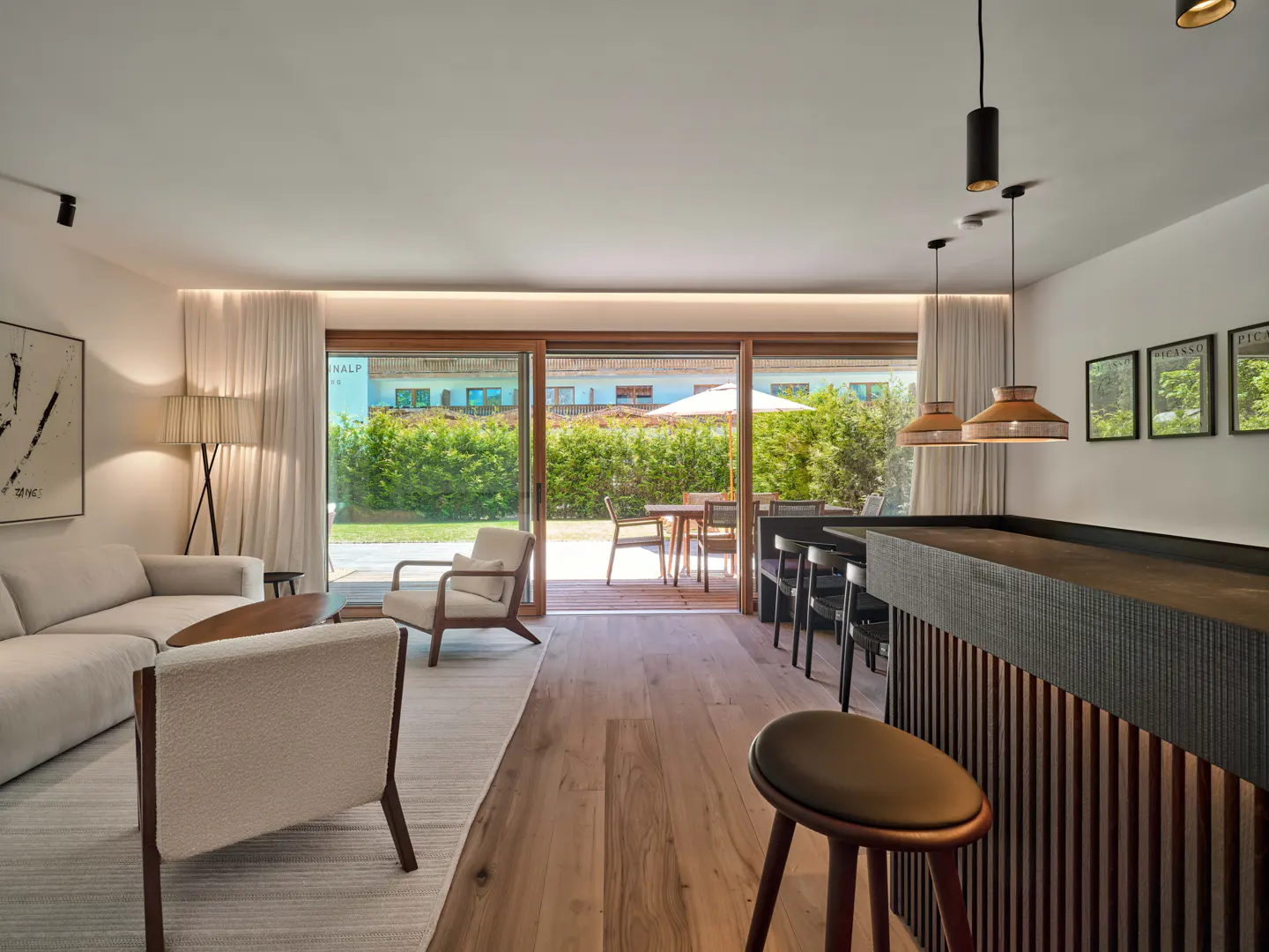 Modern living room with a white sofa, armchairs, and a wooden bar counter, opening to a patio with outdoor dining furniture.
