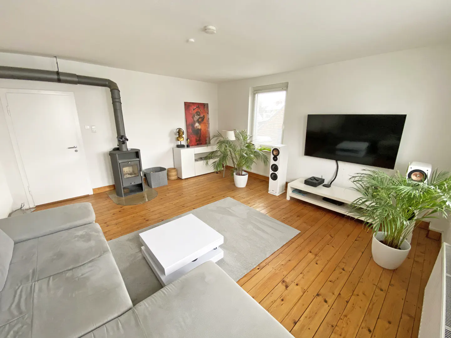 Bright living room with wood floors, a gray sectional sofa, a white coffee table, and a black fireplace. A large TV and speakers are on a white console.