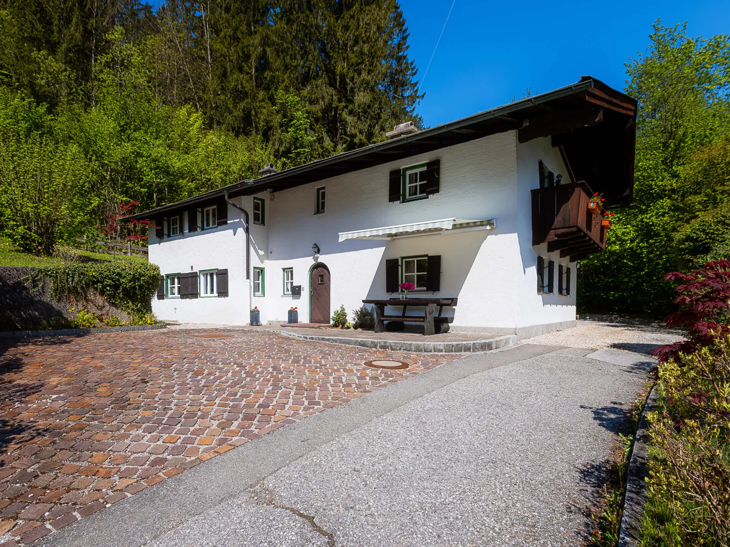 Exterior of a white two-story house with dark shutters and a cobblestone driveway, surrounded by green trees.