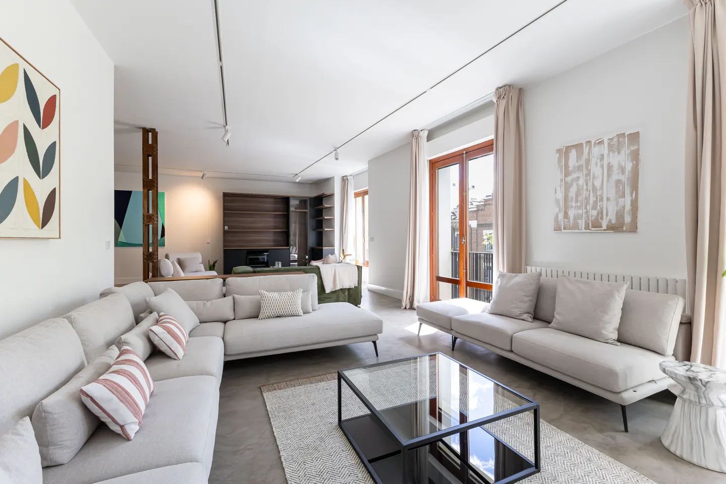 Bright, modern living room with a large sectional sofa, glass coffee table, and balcony access. Artwork adorns the white walls.